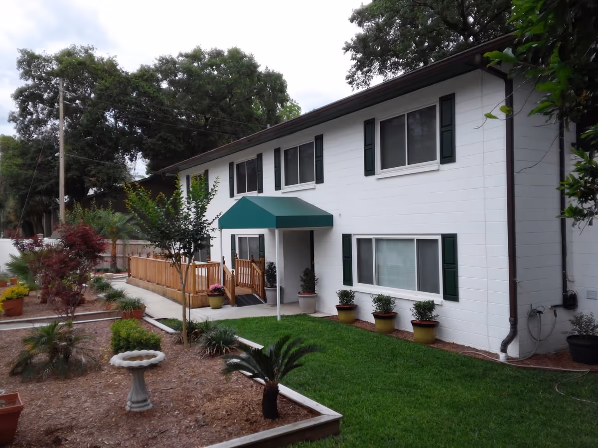 Two-story white building with green shutters and awning, front lawn, potted plants, and a wooden ramp entrance.
