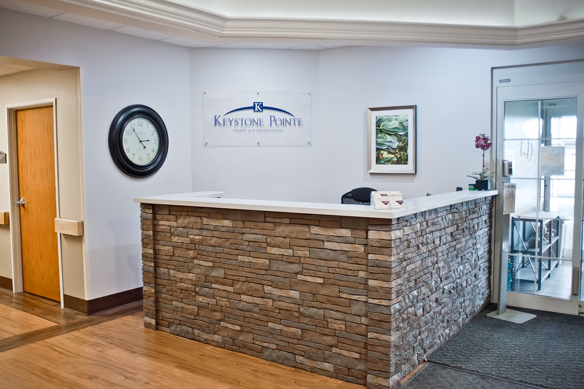 Reception desk area inside Keystone Pointe facility with a stone facade counter, a wall clock showing 11:55, a framed plant picture, and a glass door entrance on the right.