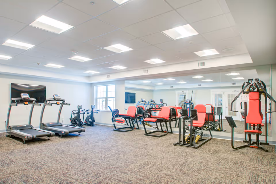 A bright and spacious fitness room with various exercise equipment including two treadmills, stationary bikes, and multiple strength training machines with red padded seats. The room has large windows, a wall-mounted TV, and a large mirror covering one wall.