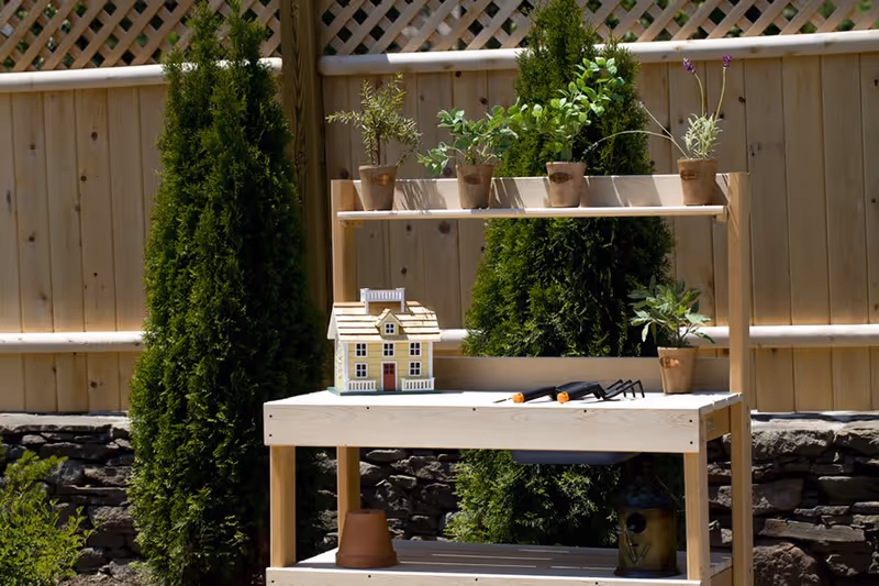 A wooden potting bench outdoors with several small potted plants on the top shelf and surface. There is a small model house and gardening tools on the bench. Behind the bench is a wooden fence and tall green shrubs.