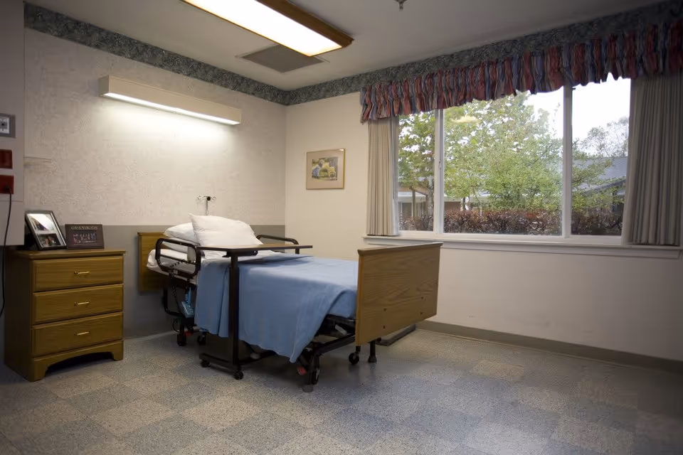 A simple patient room with a hospital bed, wooden nightstand, and a large window overlooking trees.