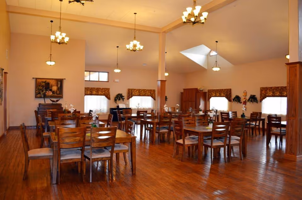 A spacious dining room with multiple wooden tables and chairs arranged neatly. The room has warm lighting from chandeliers hanging from the ceiling and large windows with floral valances allowing natural light to enter. The floor is wooden, and there are decorative plants and artwork on the walls.