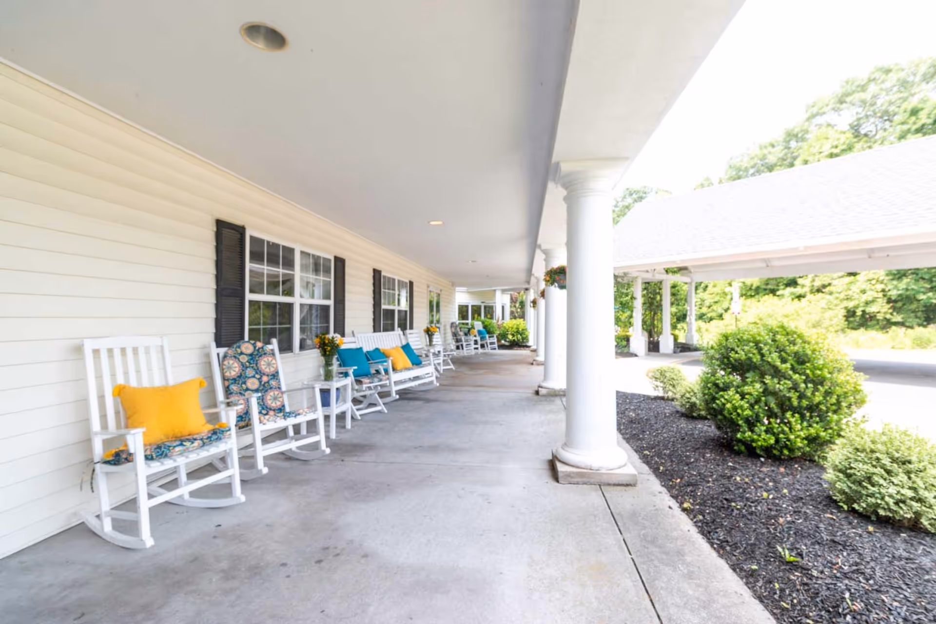 Covered front porch of a senior living facility with white rocking chairs adorned with colorful cushions, white columns, and landscaped shrubs.