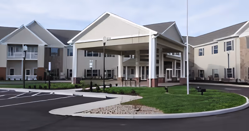 Front exterior view of a senior living facility building with a covered entrance, multiple windows, and a circular driveway with parking spaces and landscaped green areas.