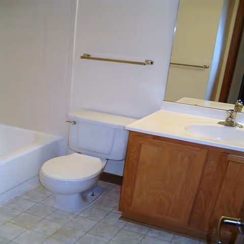 A bathroom featuring a white toilet, a bathtub with white tiled walls, a wooden vanity with a white countertop and sink, a large mirror above the sink, and a towel bar mounted on the wall above the toilet.