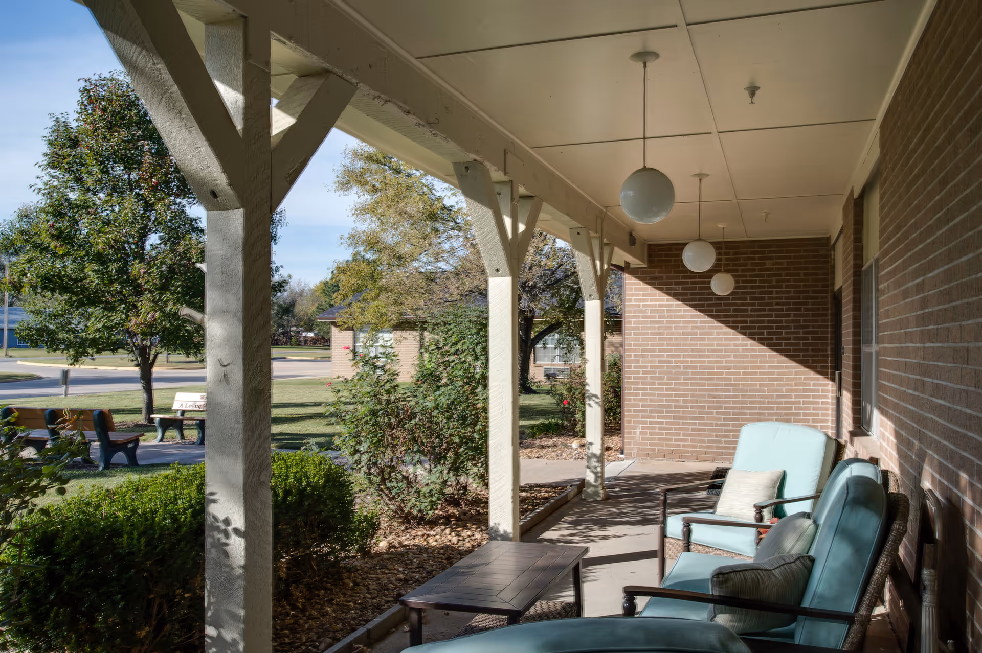 A covered outdoor patio area with cushioned chairs and a small table, overlooking a garden with bushes, trees, and benches. The patio has hanging globe lights and is adjacent to a brick building.