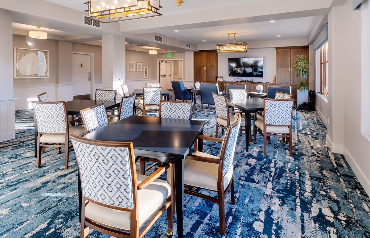 A spacious dining room with multiple dark wood tables and patterned upholstered chairs arranged neatly on a blue and white patterned carpet. The room features large windows on the right side allowing natural light to fill the space, modern rectangular ceiling light fixtures, and a large flat-screen TV mounted on a wooden cabinet against the far wall. There are also some plants and framed artwork on the walls.
