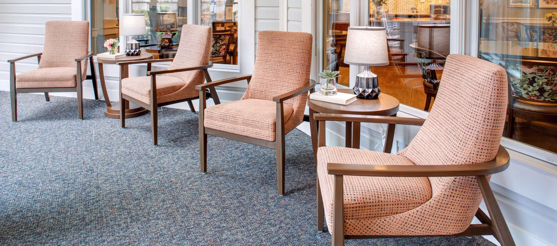 Four upholstered armchairs with side tables and lamps arranged along interior windows in a lounge.
