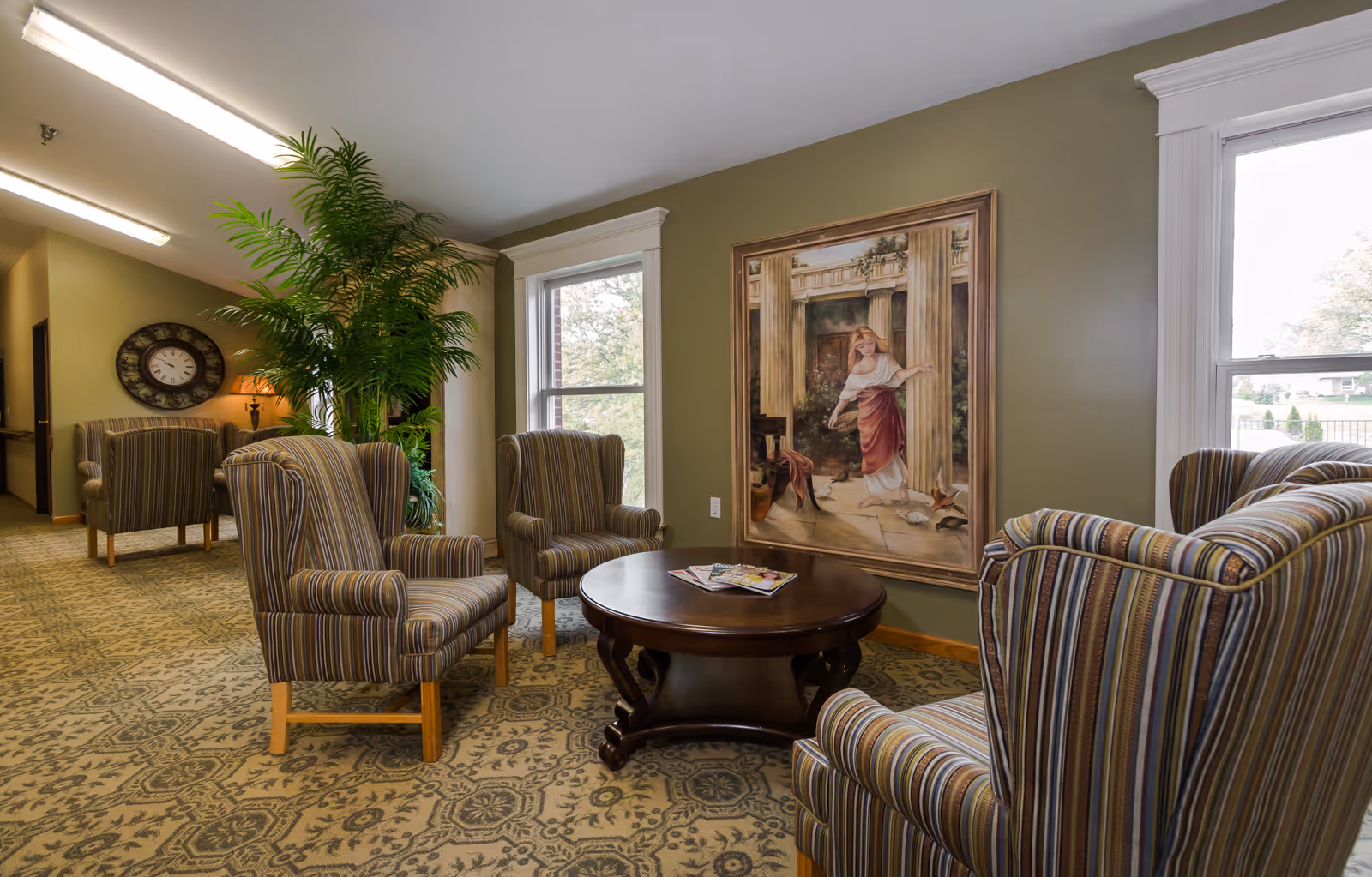 A cozy sitting area in a senior living facility with four striped armchairs arranged around a round wooden coffee table holding magazines. The walls are painted olive green and decorated with a large framed painting of a woman near classical columns. There are two large windows letting in natural light and a tall green plant near the seating area. A hallway with additional chairs and a wall clock is visible in the background.