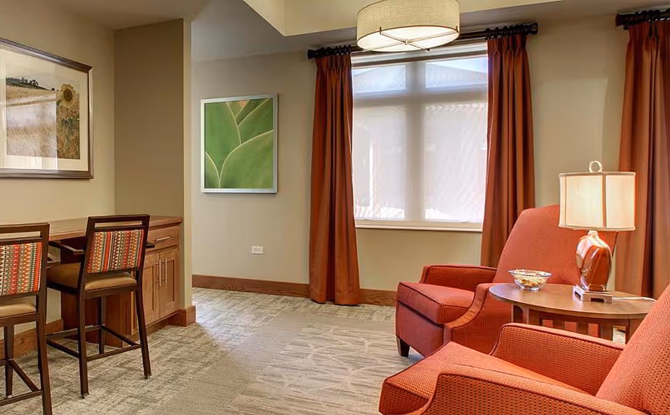 A cozy senior living room with two red upholstered armchairs, a round wooden side table with a lamp and a bowl, a window with red curtains, a ceiling light fixture, and a small wooden desk with two chairs featuring patterned cushions. The walls are decorated with framed artwork.