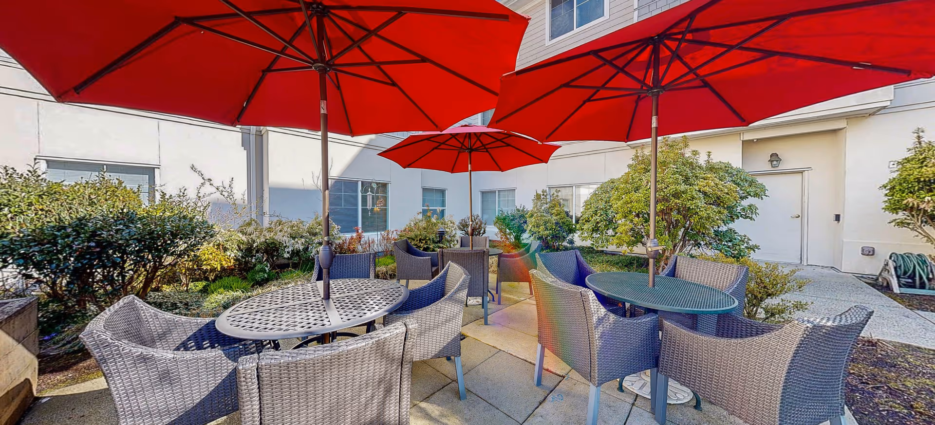Outdoor patio area with multiple round tables and wicker chairs under large red umbrellas, surrounded by bushes and plants, adjacent to a building with windows and a door.