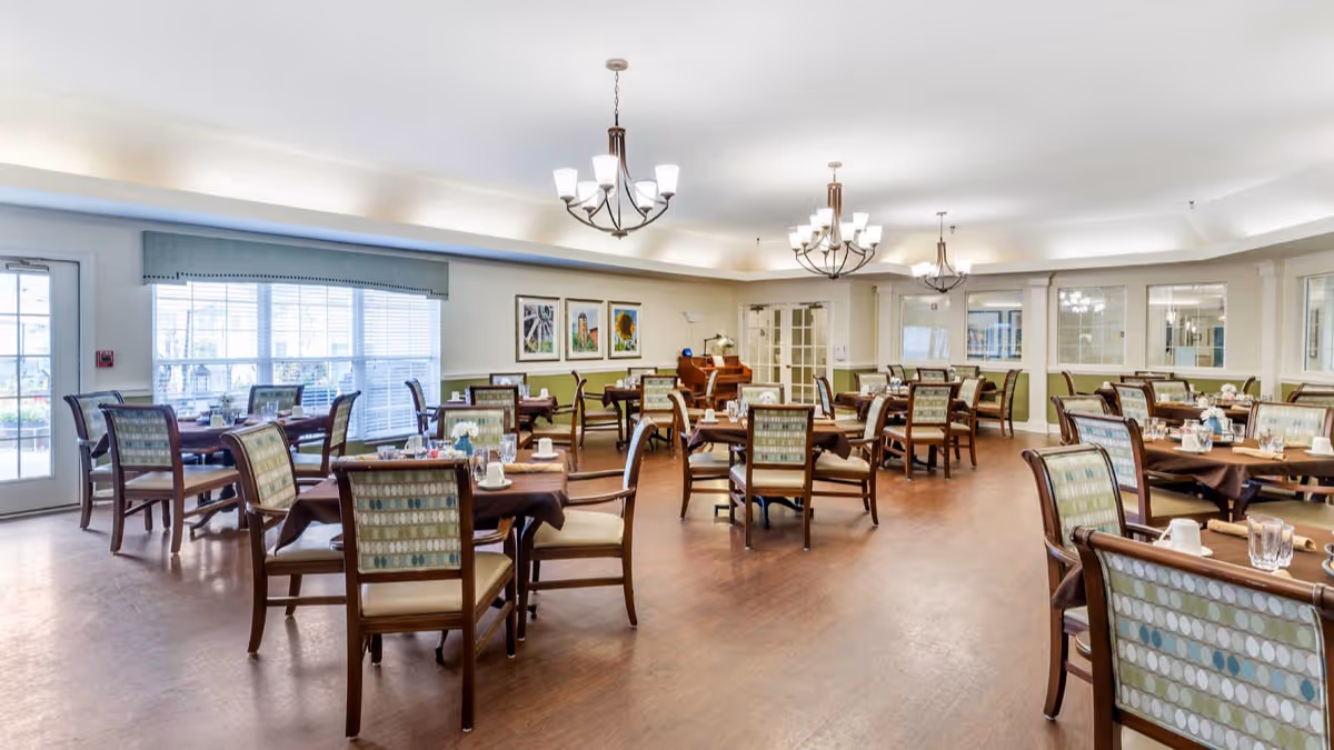 A spacious, well-lit dining room with multiple wooden tables and chairs arranged neatly. Each table is set with cups, glasses, and napkins. The room features large windows with blinds, three chandeliers hanging from the ceiling, framed artwork on the walls, and a piano in the background.