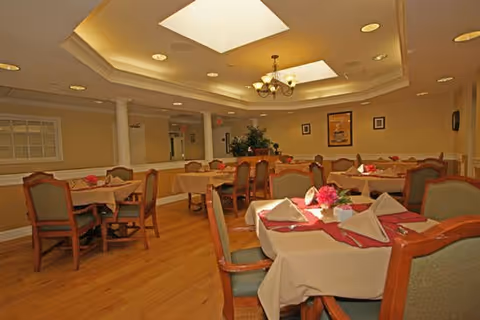 Well-lit dining room with multiple tables set with napkins and floral centerpieces, wooden chairs, chandeliers, and skylights.