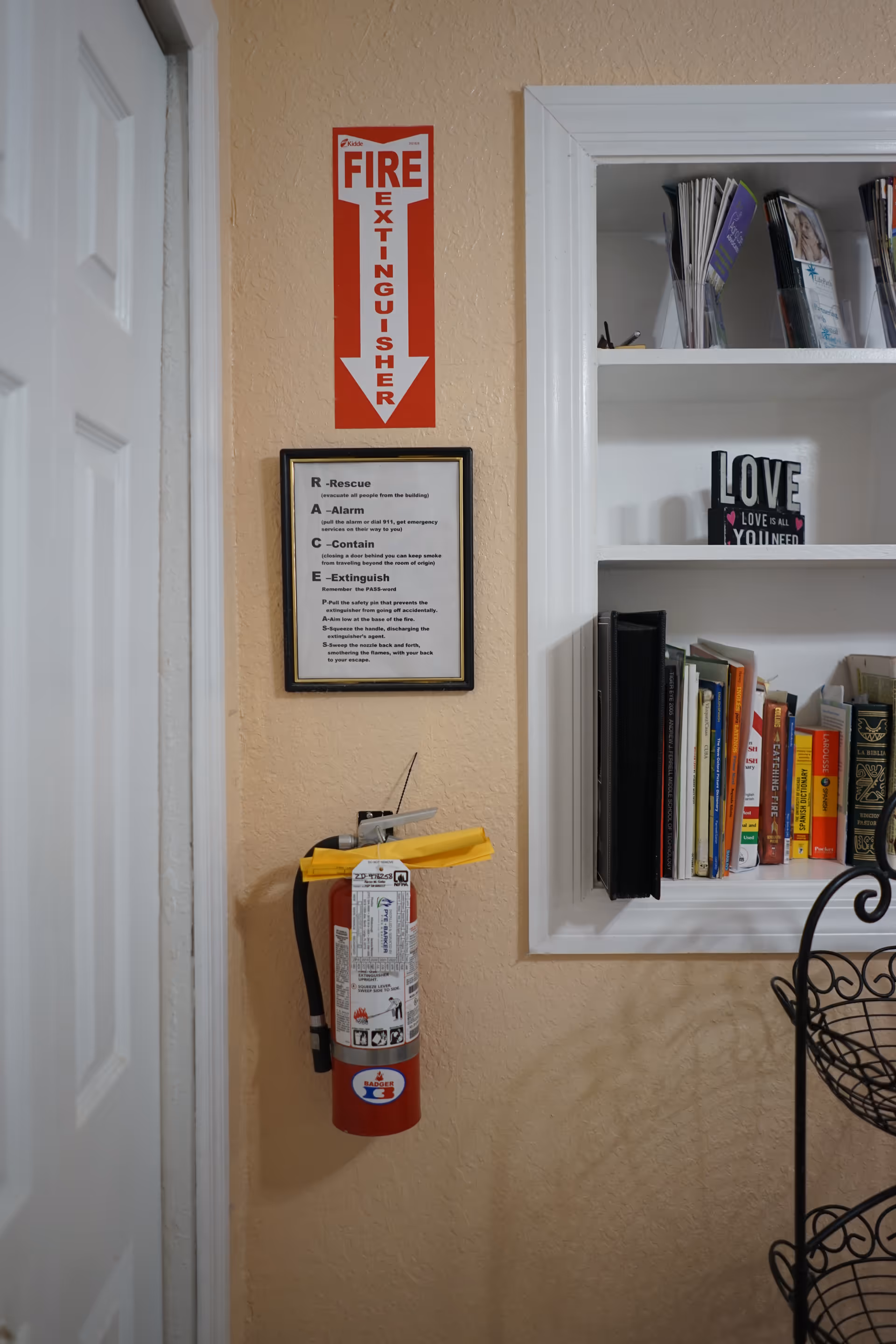 A fire extinguisher mounted on a beige wall below a red and white sign with a downward arrow labeled 'FIRE EXTINGUISHER'. Above the extinguisher is a framed instructional sign with steps for using the fire extinguisher. To the right is a built-in white bookshelf containing books and decorative items, including a block sign that says 'LOVE'. A white door is partially visible on the left side of the image.