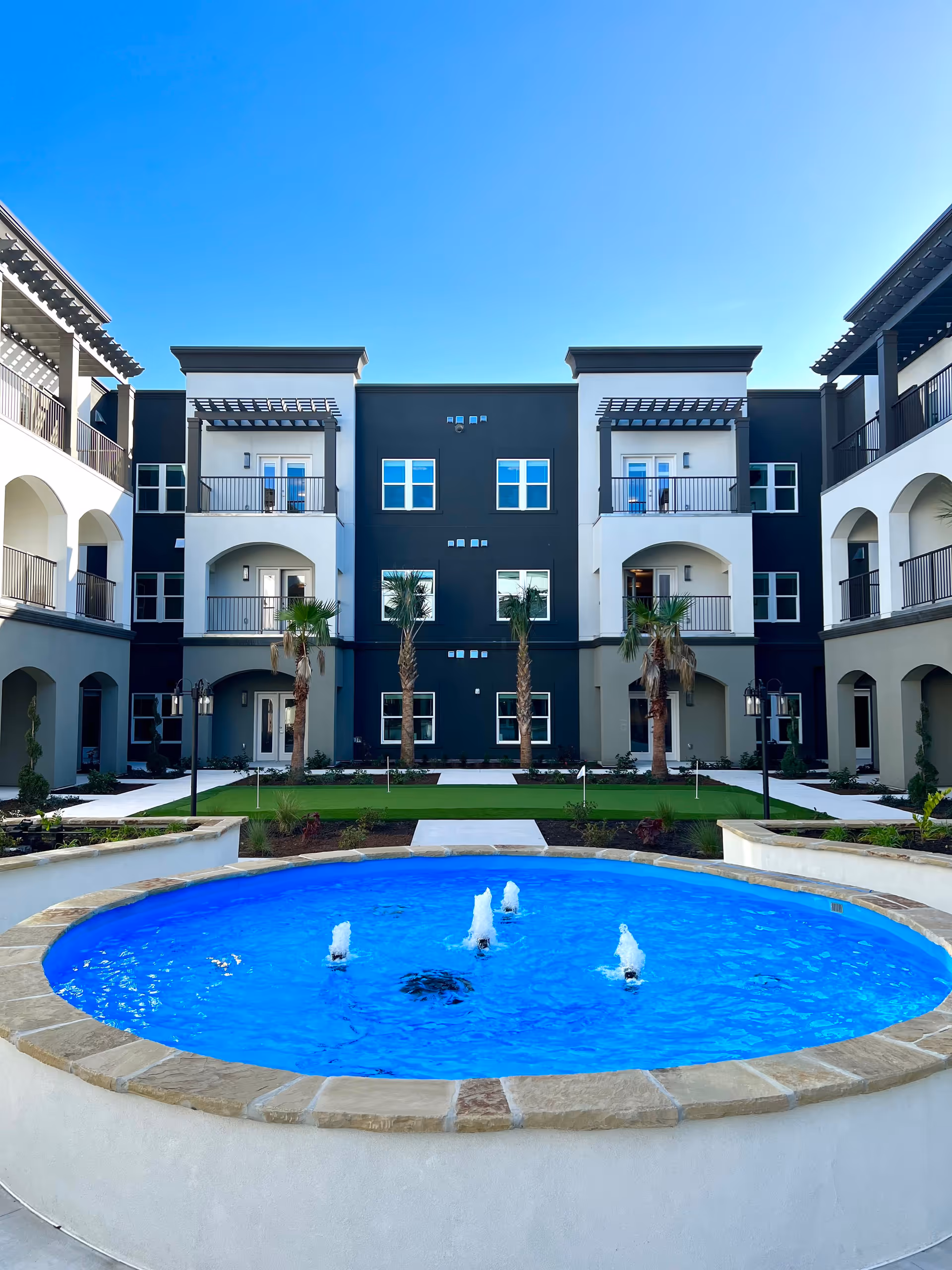 A bright blue fountain in a courtyard in front of a three-story building with balconies, palm trees, and a clear sky.