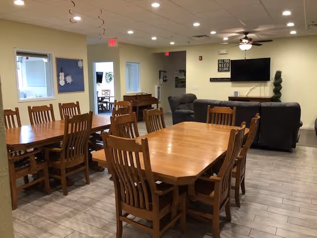 A dining area with multiple wooden tables and chairs arranged neatly. In the background, there is a living room area with a dark gray sofa and armchair facing a wall-mounted TV. The walls are light-colored, and the floor has light gray tiles. Ceiling lights and a ceiling fan are visible, along with some wall decorations and an exit sign.