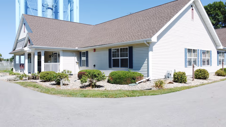 Exterior view of a single-story building with white siding and a brown shingled roof, surrounded by small bushes and landscaping with rocks. The building has multiple windows with blue shutters and a small covered porch area. The sky is clear and blue.