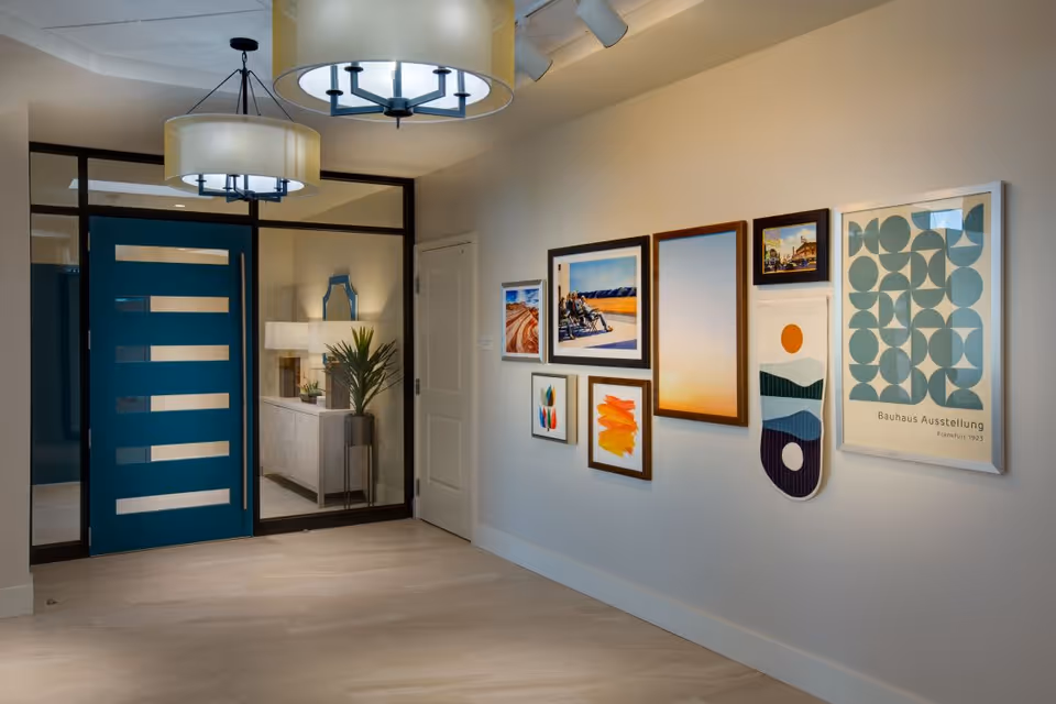 Interior hallway of a senior living facility with a modern blue door and glass panels. The hallway features light wood flooring, two large ceiling light fixtures, and a wall decorated with various framed artworks and prints. A console table with a lamp and a plant is visible through the glass panel next to the door.