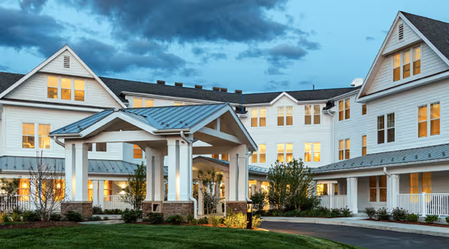 Exterior view of a large, white senior living facility building at dusk with multiple windows lit from inside. The building has a covered entrance with white pillars and a metal roof, surrounded by landscaped greenery and a paved driveway.