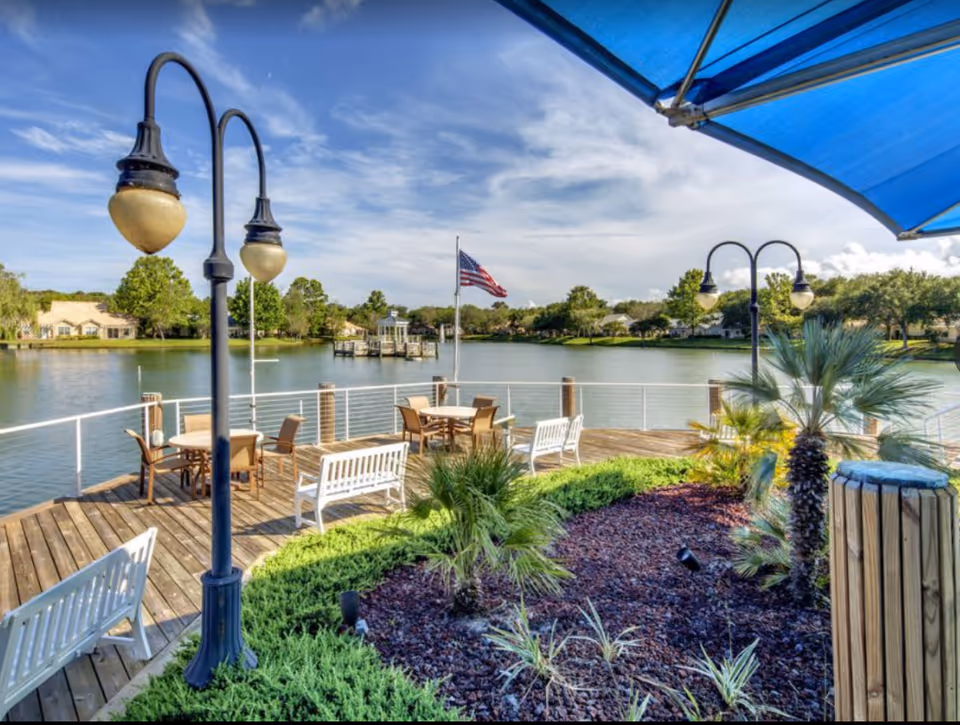 Lakeside wooden deck with benches, tables, lamp posts, an American flag, and landscaped plants under a blue sky.