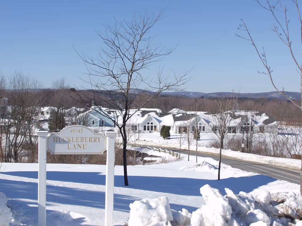 Snow-covered landscape with a road leading to a group of white buildings in the distance under a clear blue sky. A sign in the foreground reads '40-43 HUCKLEBERRY LANE'. Leafless trees are scattered around the area.