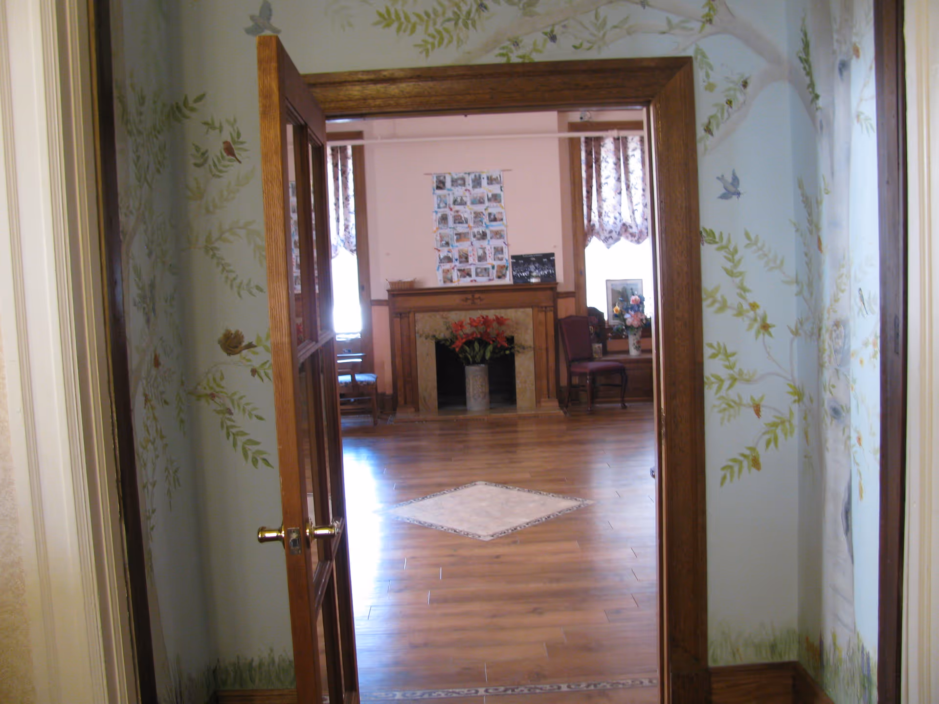 View through a wooden door with glass panels into a room with wooden flooring, a fireplace decorated with flowers, chairs, and windows with floral curtains. The walls near the door have a nature-themed mural with birds and leaves.