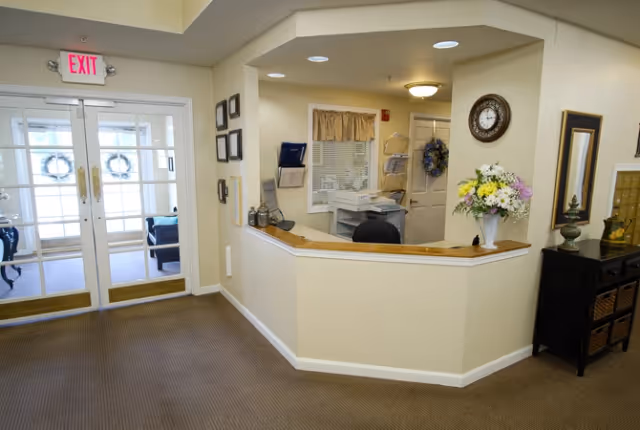 Reception area inside Cedar Creek of Bedford facility with a front desk, a computer, a clock on the wall, a vase of flowers, and double glass doors leading outside with exit signs above.