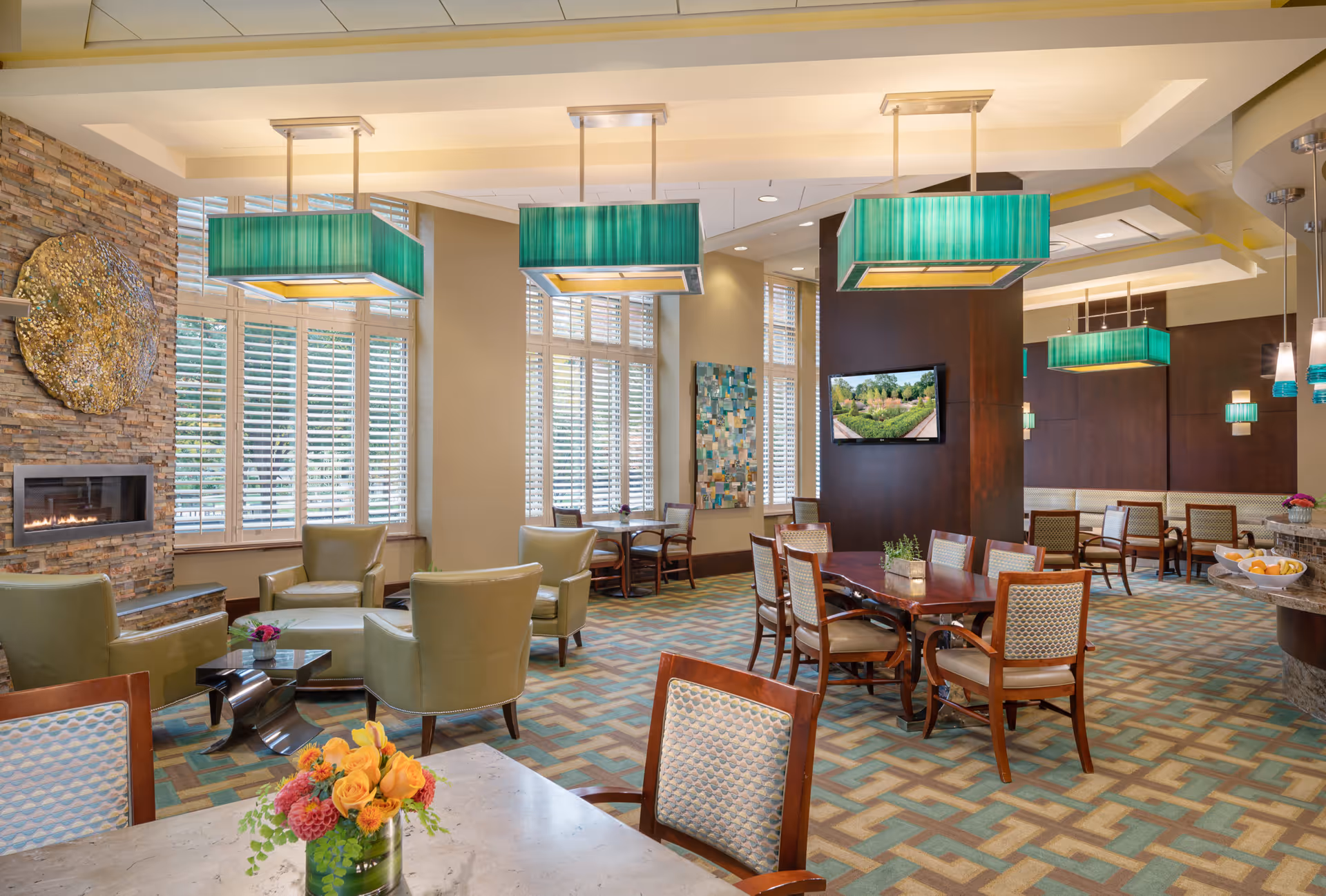 Bright communal dining and lounge area with tables and chairs, green pendant lights, a stone fireplace, and a wall-mounted TV.