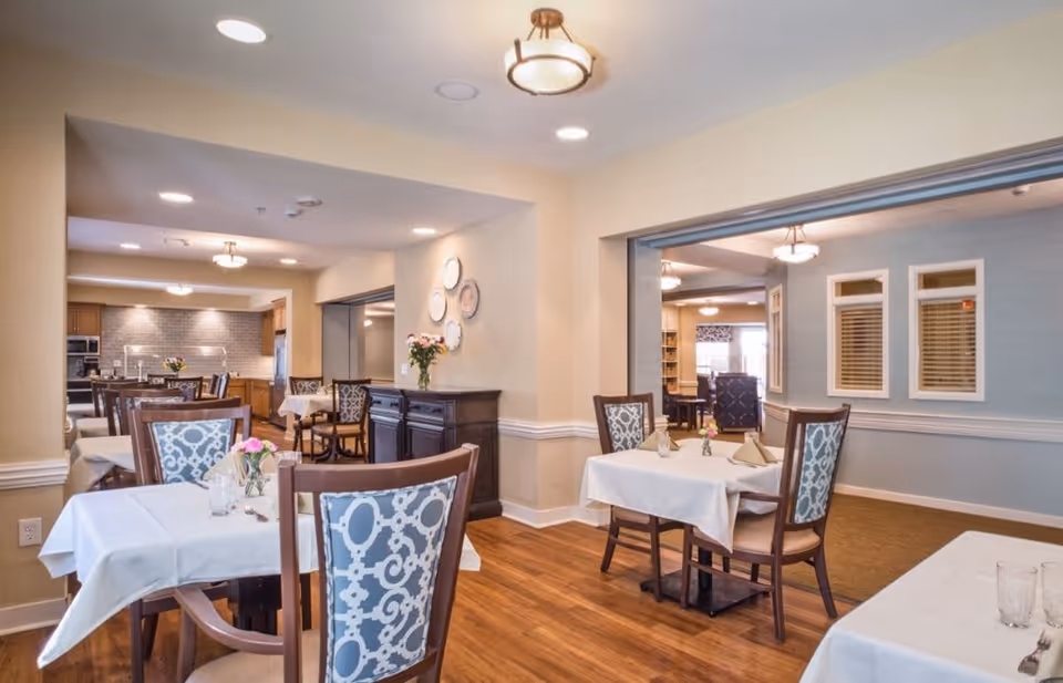 A bright dining room in a senior living facility with several tables covered in white tablecloths, each set with glasses, napkins, and small flower vases. The room features wooden floors, patterned dining chairs, and soft lighting from ceiling fixtures. There is an open view into an adjacent room with more seating and a kitchen area in the background.