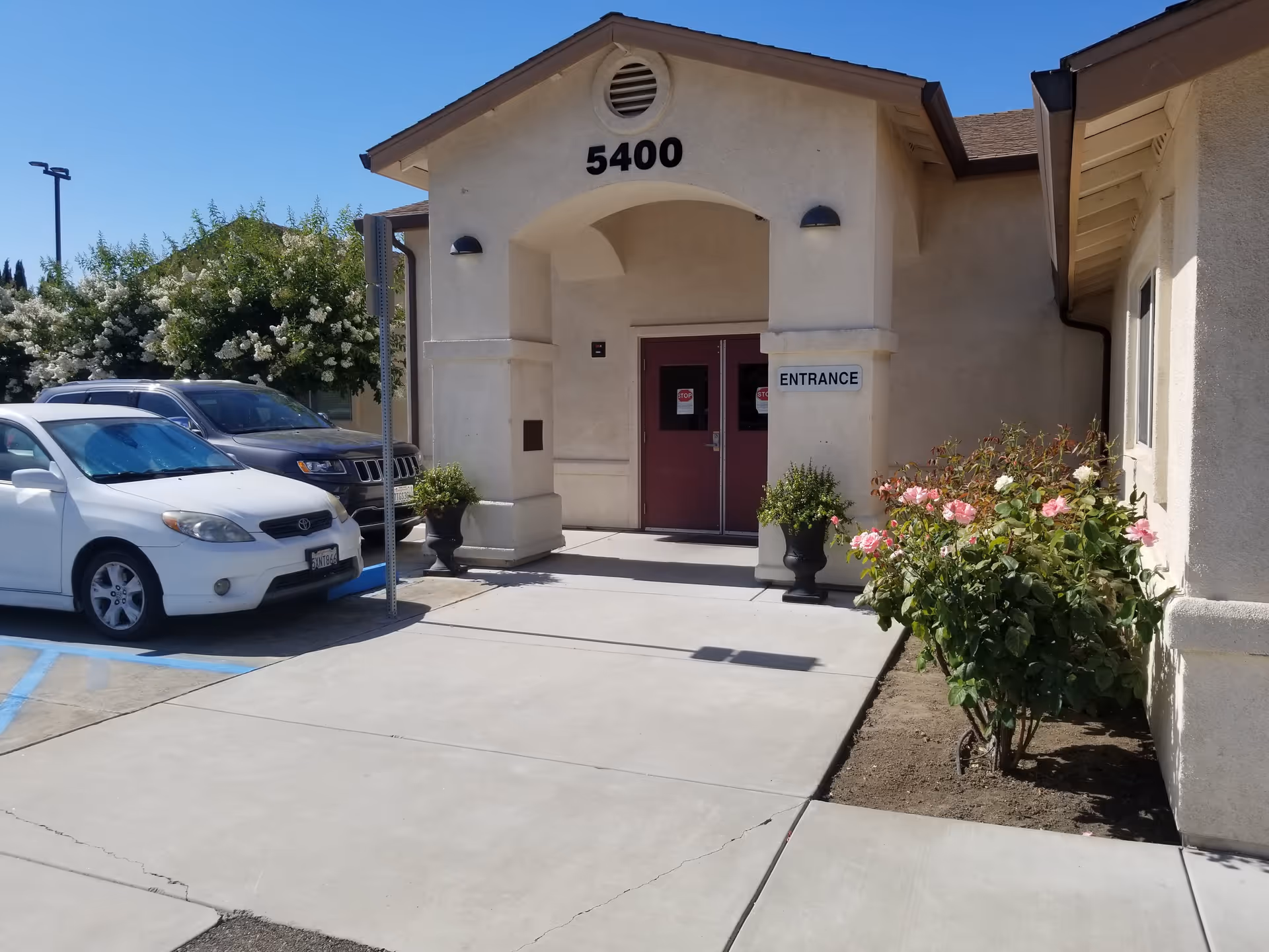 Exterior view of a building entrance with the number 5400 above the archway. There are two maroon double doors with 'STOP' signs on them and a white sign that says 'ENTRANCE' on the right pillar. Two black planters with green plants flank the entrance. To the right, there is a flower bed with blooming pink and white roses. On the left side, there are parked cars and some trees with white flowers in the background under a clear blue sky.
