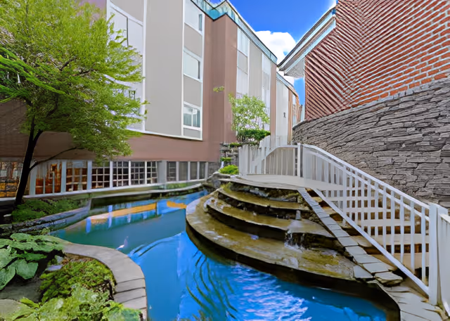 Courtyard with a tiered stone water feature and pond crossed by a white wooden footbridge between multi-story buildings.