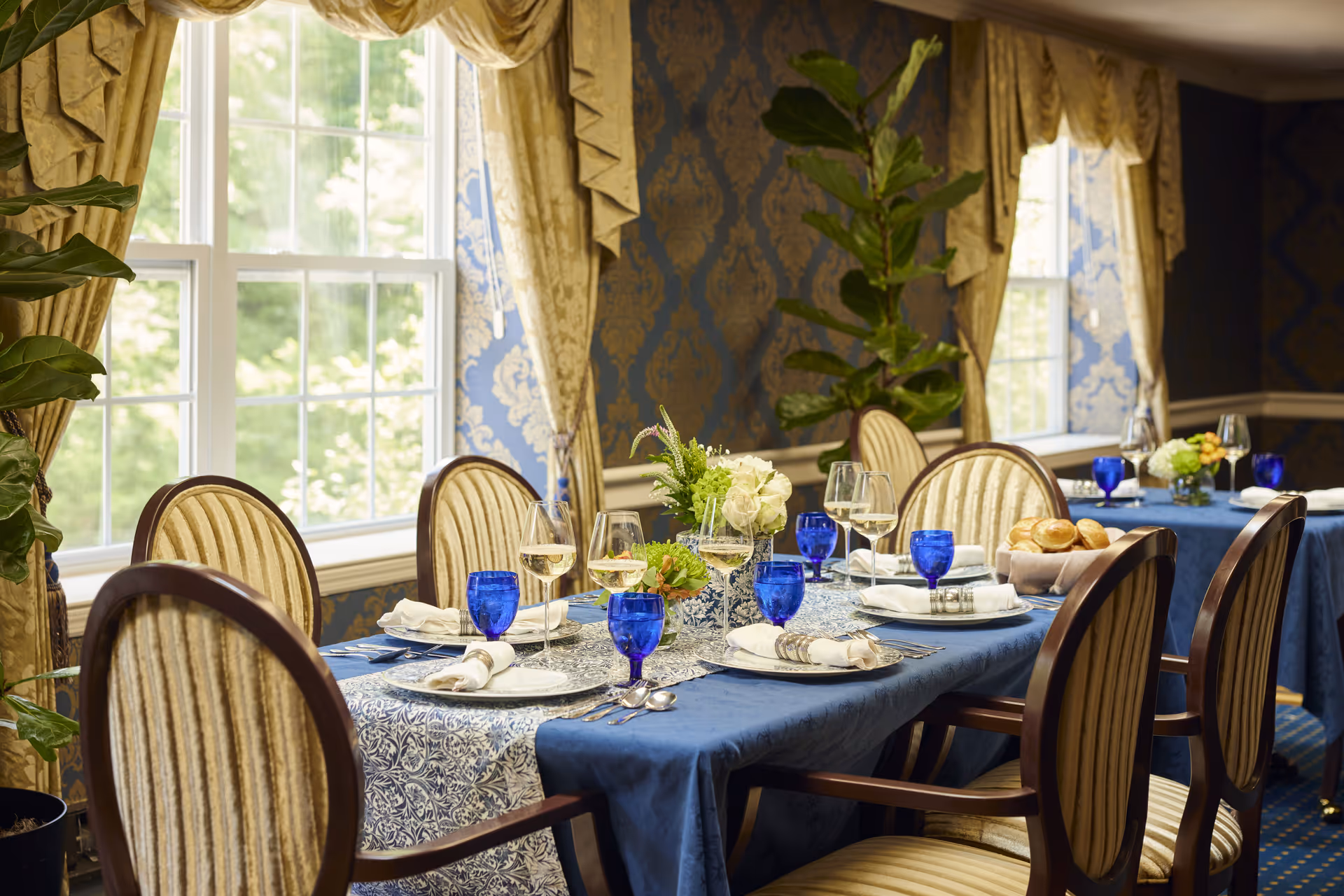 A formal dining room with a table set for a meal. The table is covered with a blue tablecloth and a patterned runner, and is set with white plates, silverware, white napkins with napkin rings, blue water glasses, and wine glasses filled with white wine. There are small floral centerpieces and a basket of bread rolls on the table. The room features large windows with gold drapes, blue and gold patterned wallpaper, and upholstered dining chairs with wooden frames.