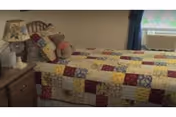 Single bedroom with a twin bed covered in a patchwork quilt, a nightstand holding a lamp and stuffed animals, and a window with an air conditioner.