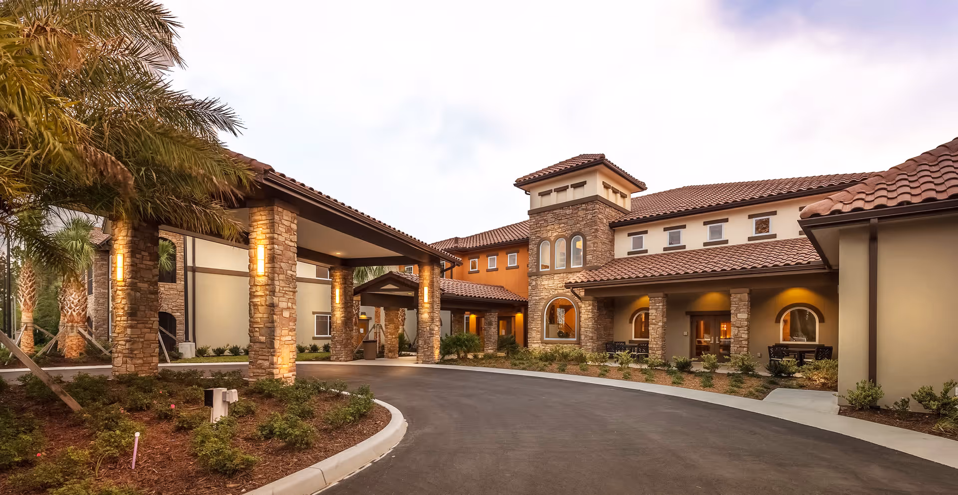 Exterior view of HarborChase of Palm Coast senior living facility featuring a curved driveway, stone pillars, and a building with a tiled roof and arched windows, surrounded by landscaped greenery and palm trees under a partly cloudy sky.