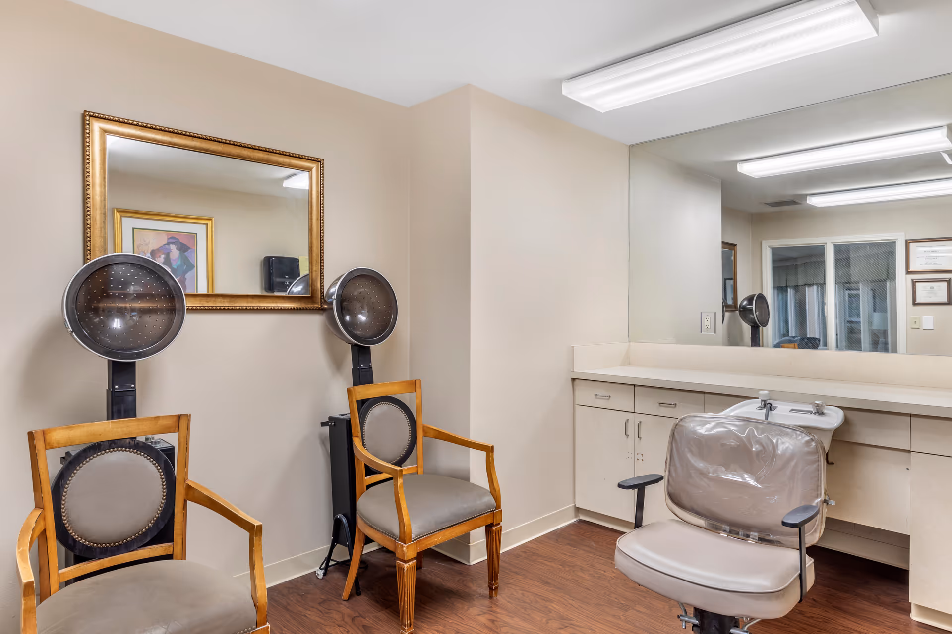 Interior of a salon area in a senior living facility with two vintage hair dryer chairs against a beige wall, a large gold-framed mirror above them, and a salon chair with a plastic cover in front of a counter with cabinets and a large wall mirror.