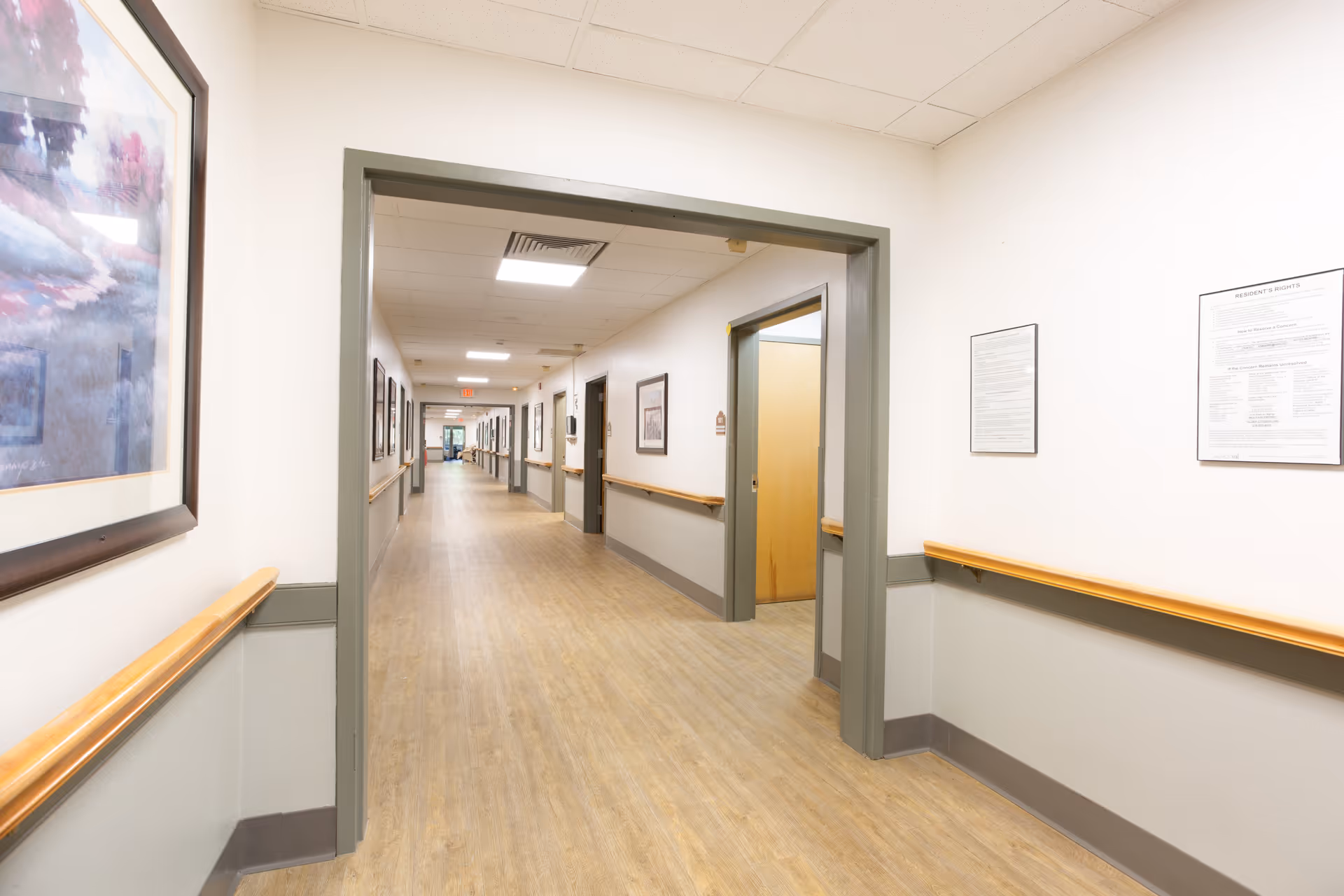 A long, well-lit hallway in a senior living facility with light wood flooring, white walls with gray trim, handrails on both sides, framed artwork and notices on the walls, and several doorways leading to other rooms.