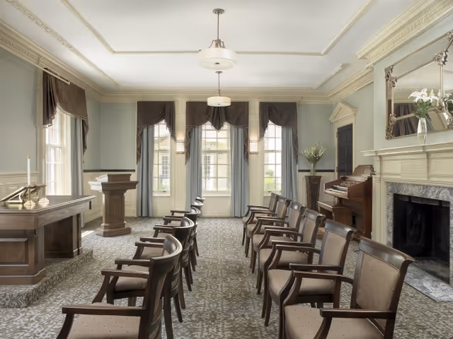 A formal room with rows of wooden chairs arranged facing a wooden podium. The room features large windows with draped curtains, a fireplace with a large mirror above it, a piano, and a wooden table with candles. The walls are painted light gray with white trim and molding, and the floor is covered with a patterned carpet.