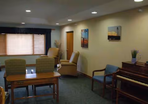 A cozy senior living lounge featuring chairs, a piano, a table, window blinds, and wall art.