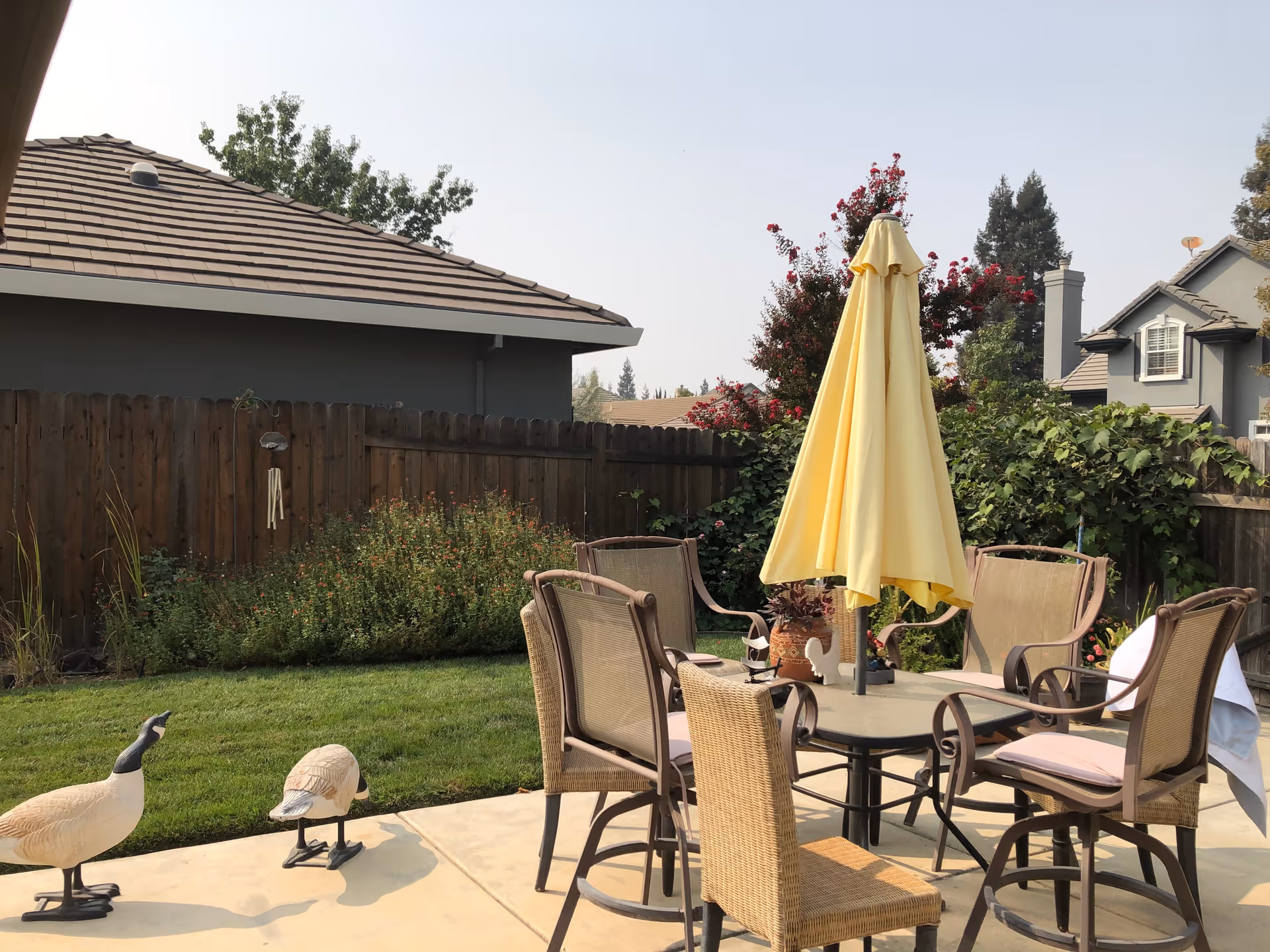 Outdoor patio area with a round table surrounded by six chairs, one of which is a wicker chair. A closed yellow patio umbrella is in the center of the table. The patio is adjacent to a grassy yard with bushes and a wooden fence. Two decorative bird statues are placed on the patio near the grass. Houses and trees are visible in the background under a clear sky.