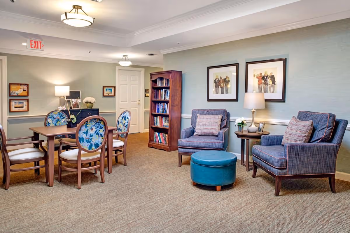 A cozy common area in a senior living facility featuring two blue upholstered armchairs with patterned pillows, a round wooden side table with a lamp and flowers, a blue ottoman, a wooden bookshelf filled with books, and a wooden table with four chairs that have floral patterned backs. The walls are light green with white trim, decorated with framed artwork, and there is a door and an exit sign in the background.