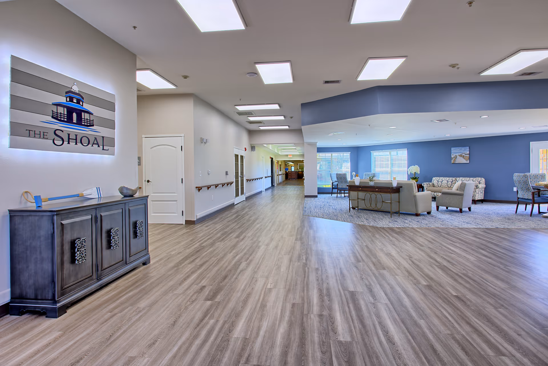 Interior view of The Shoal senior living facility showing a wide hallway with wood flooring leading to a seating area with chairs, sofas, and tables. The walls are painted in neutral and blue tones, and there is a sign on the left wall with The Shoal logo. The space is well-lit with ceiling lights and natural light from windows.