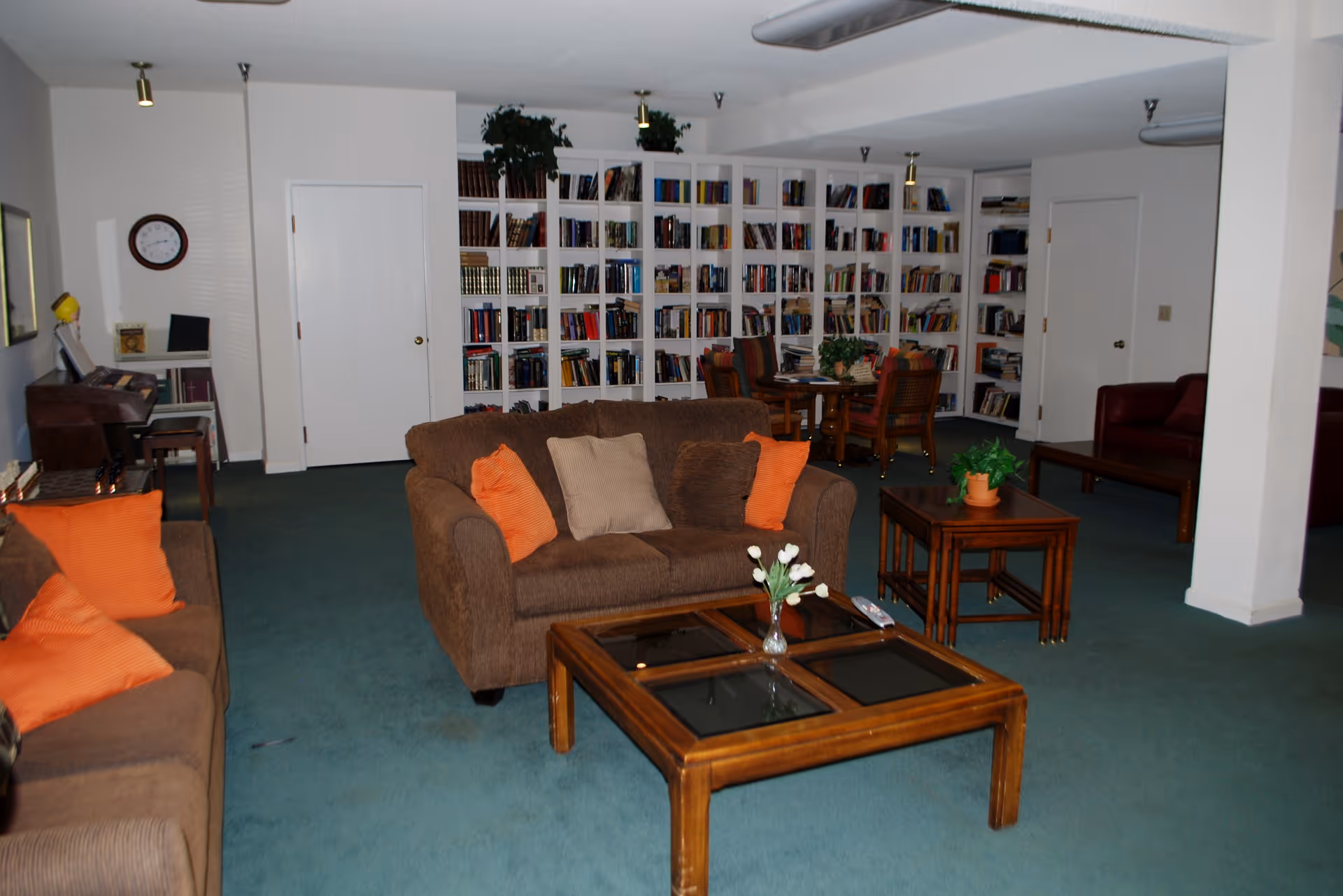 A cozy living room area with brown sofas adorned with orange and beige cushions, a wooden coffee table with a vase of white flowers, and a side table with a potted plant. In the background, there is a large white bookshelf filled with books, a round table with chairs, and a piano against the wall. The room has green carpet and white walls with two closed white doors.