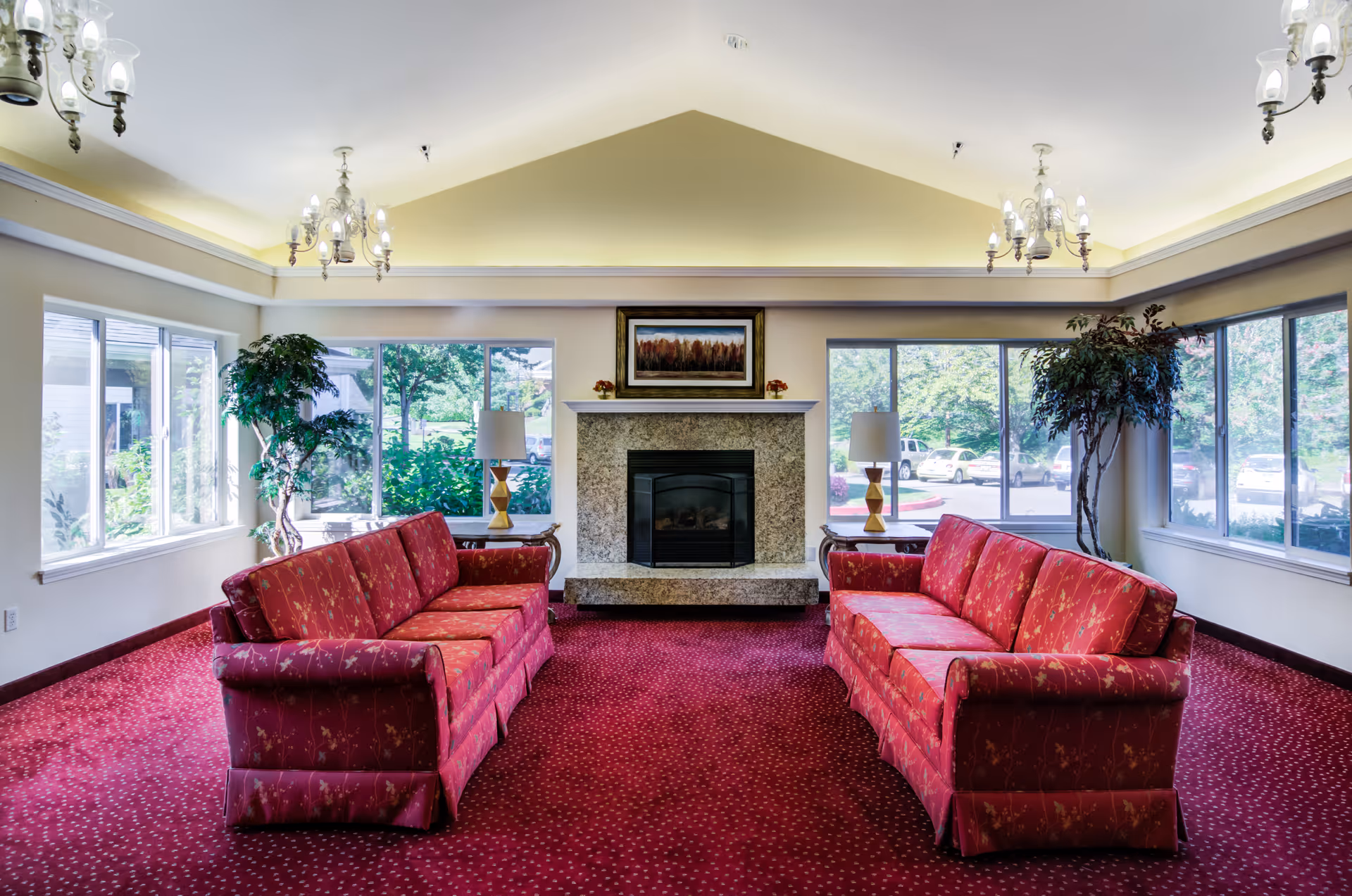 Bright communal lounge with two red sofas facing a central fireplace, large windows, potted plants and chandeliers.