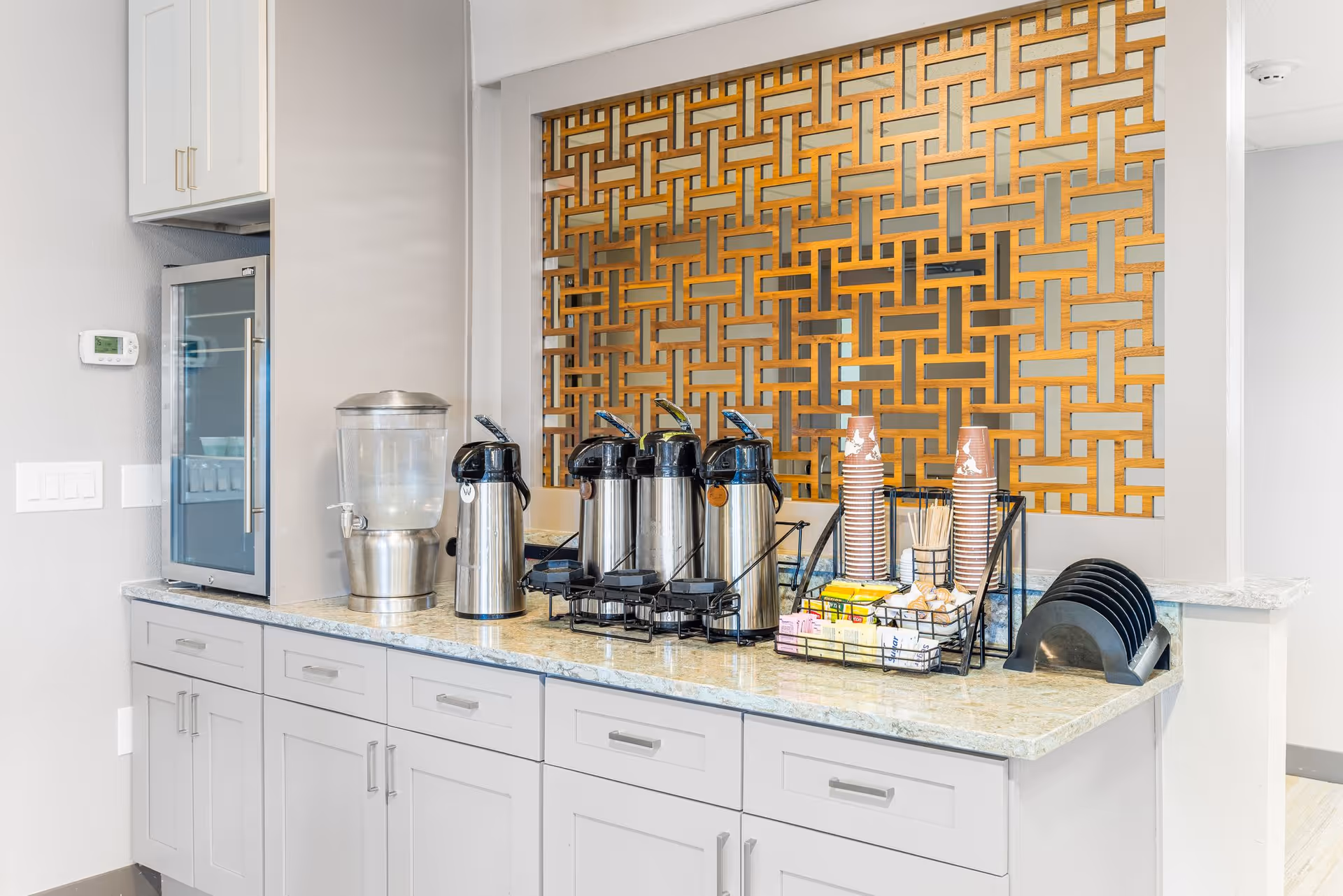 A beverage station with a water dispenser, four stainless steel coffee airpots, stacks of disposable cups, stirrers, sugar packets, and a black tray holder on a granite countertop. The background features a decorative wooden lattice panel.
