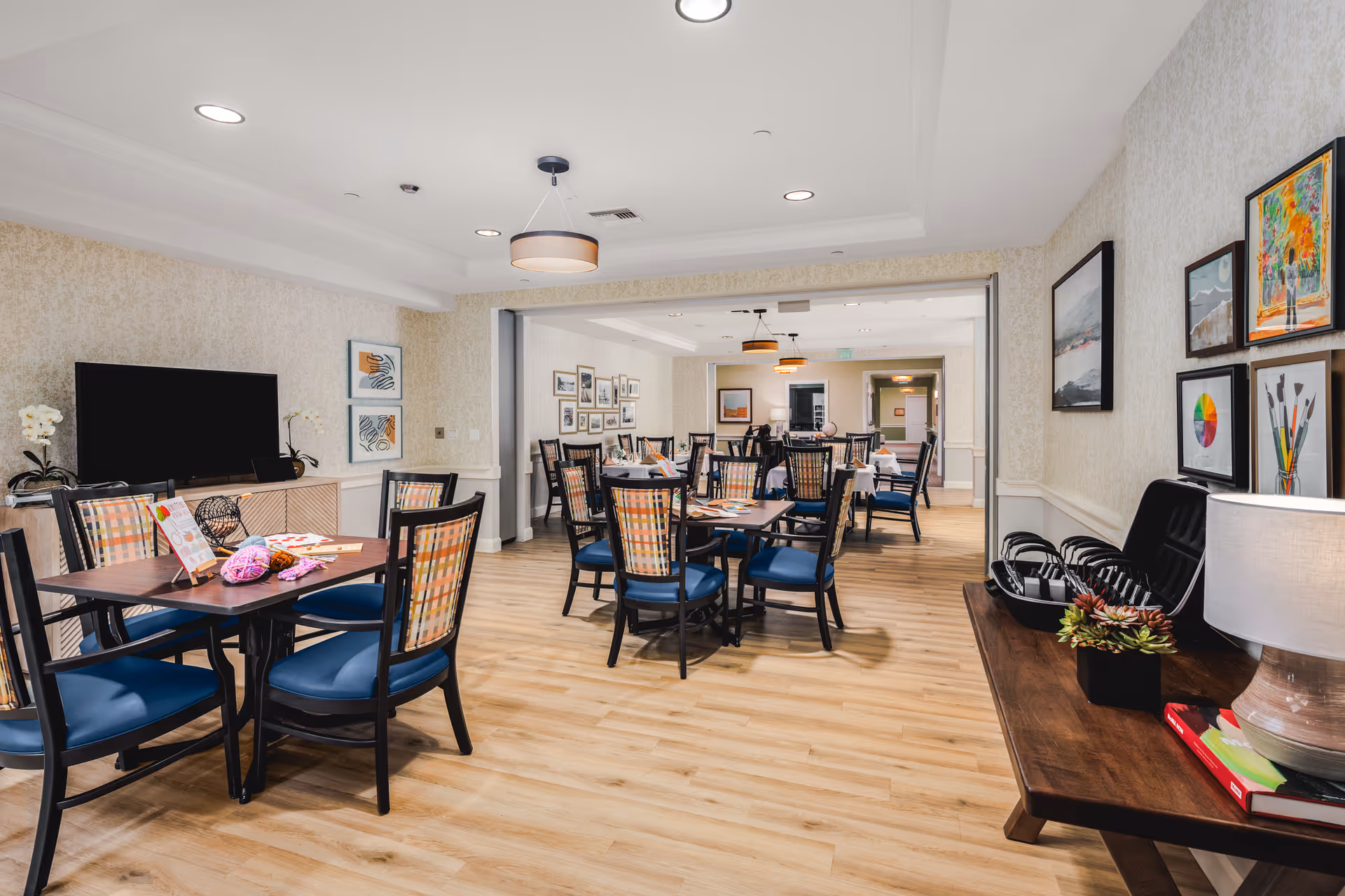 A spacious and well-lit common area in a senior living facility with multiple tables and chairs arranged for group activities or dining. The room features light wood flooring, patterned wallpaper, and framed artwork on the walls. On the left side, there is a TV on a cabinet with decorative plants, and on the right, a wooden console table with a lamp, books, and a plant. The tables have various items on them, including yarn and game pieces, suggesting recreational activities.