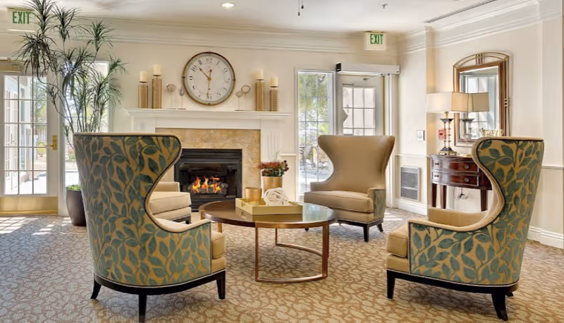 A cozy sitting area in an assisted living facility featuring a round wooden coffee table surrounded by four upholstered armchairs with leaf-patterned fabric. Behind the seating area is a fireplace with a large clock above it and decorative candles on the mantel. The room has large windows and glass doors letting in natural light, with an exit sign visible above the doors. A wooden side table with a lamp and mirror is positioned against the wall.