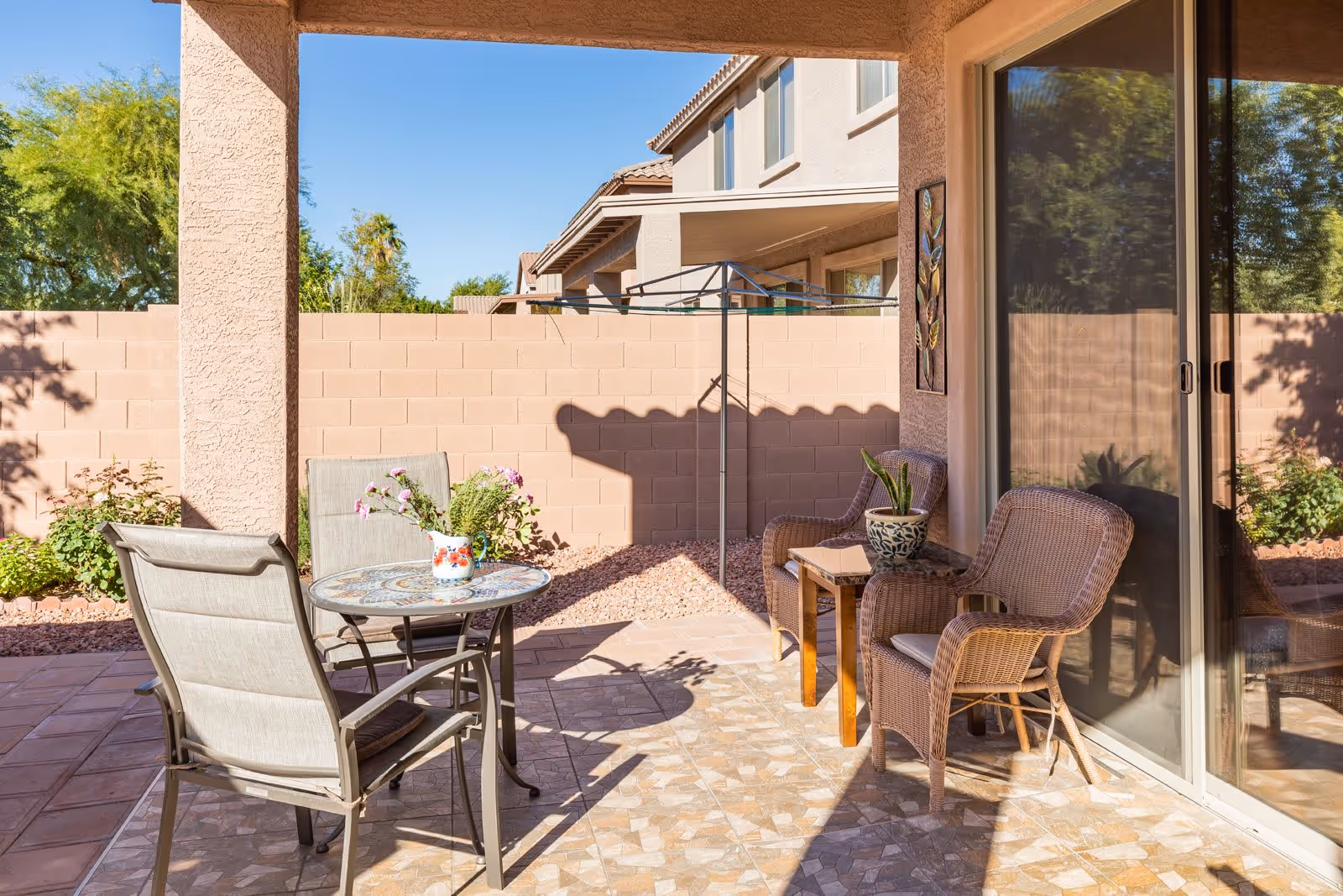 A sunny outdoor patio area with a tiled floor, featuring a round table with a floral vase and two chairs on the left, and two wicker chairs with a small wooden table and a potted plant on the right. The patio is enclosed by a beige brick wall, with a house and trees visible in the background.