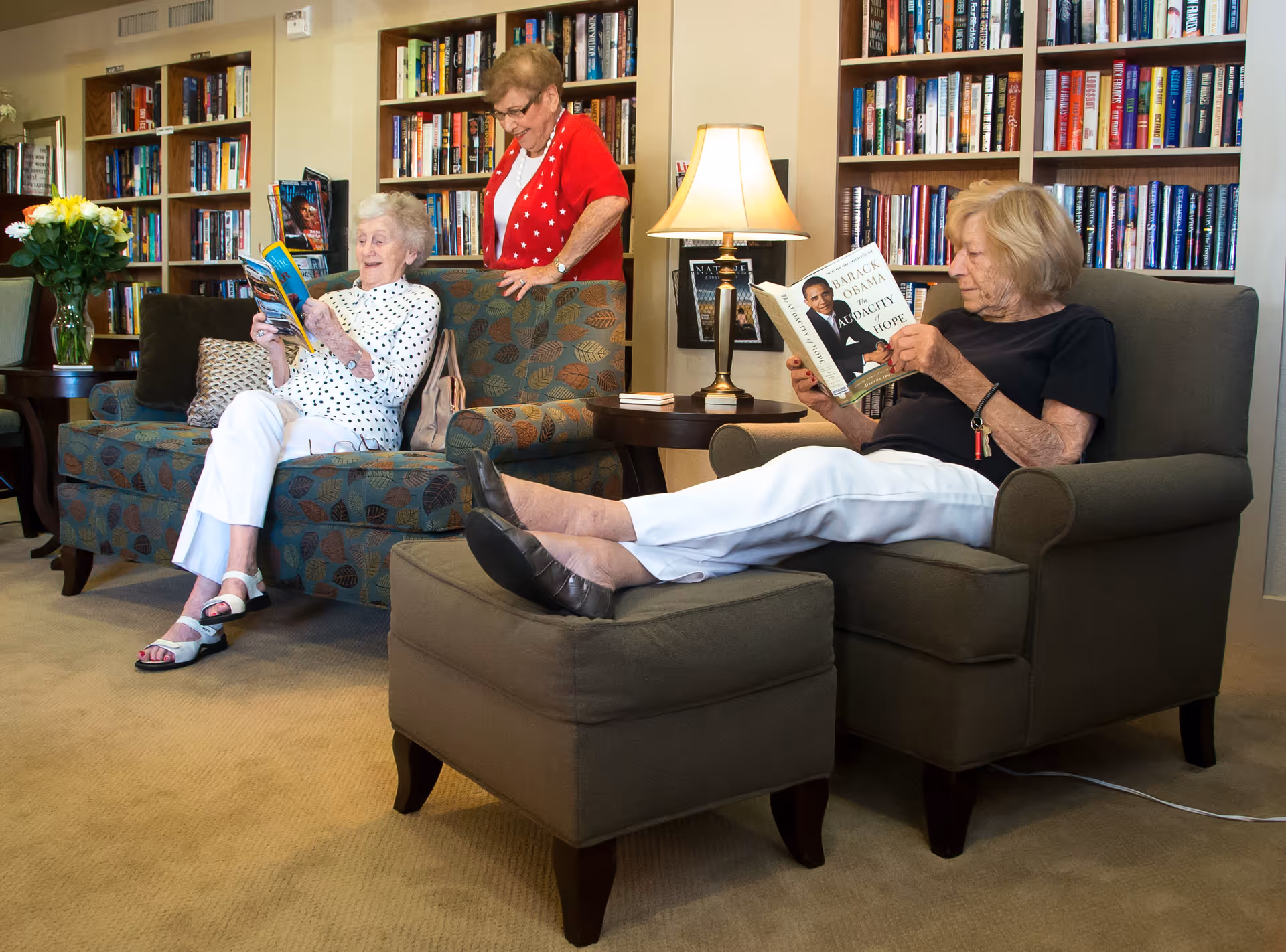 Three elderly women in a cozy library or reading room. One woman is sitting on a patterned sofa reading a magazine, another woman stands behind the sofa smiling, and the third woman is seated in an armchair with her feet up on an ottoman, reading a book titled 'The Audacity of Hope' by Barack Obama. Bookshelves filled with books line the walls, and a table lamp provides warm lighting.
