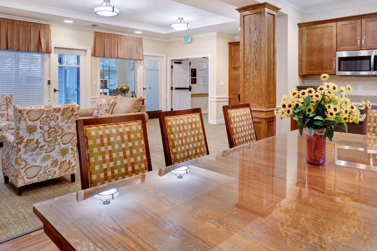 Interior view of a senior living facility showing a polished wooden dining table with a vase of sunflowers, surrounded by chairs with checkered upholstery. In the background, there is a cozy seating area with floral patterned armchairs and a beige sofa, wooden cabinetry, and a kitchen area with a microwave. The space is well-lit with ceiling lights and has a warm, inviting atmosphere.