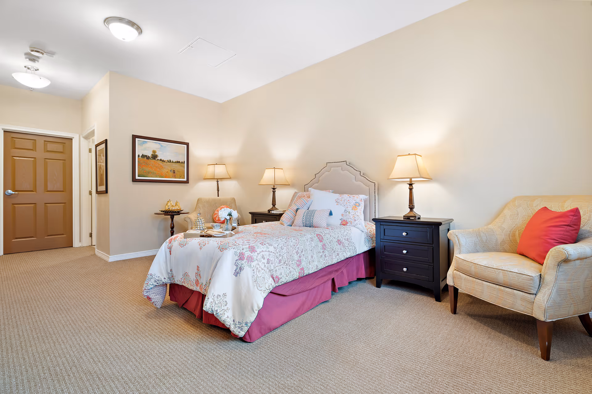 A cozy bedroom with a single bed covered in floral bedding and multiple pillows. There are two dark wooden nightstands on either side of the bed, each with a lamp. To the right of the bed is a beige upholstered armchair with a red pillow. The room has beige walls, carpeted floor, and a closed brown door in the background. A framed landscape painting hangs on the wall near the door.